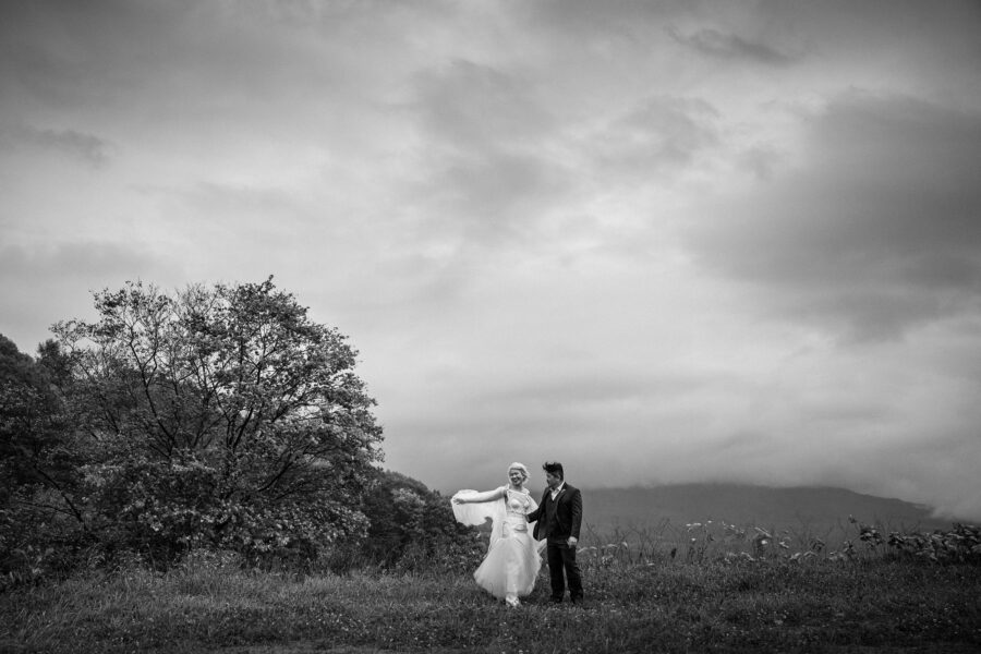 Black and white: the couple small in an open field under a stormy sky, her cape blown sideways, on their Hokkaido wedding day.