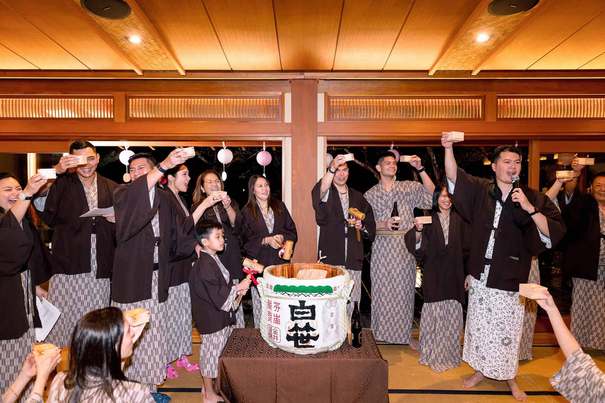 A whole family in yukata raises cups around an opened sake barrel after kagami biraki, at a ryokan welcome dinner for a wedding in Japan.