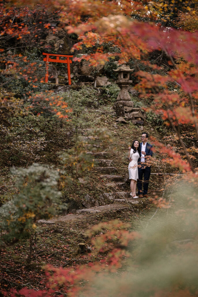 The couple stand on old stone steps below a red torii and a stone lantern, framed by autumn maples during their couple shoot in Kyoto.