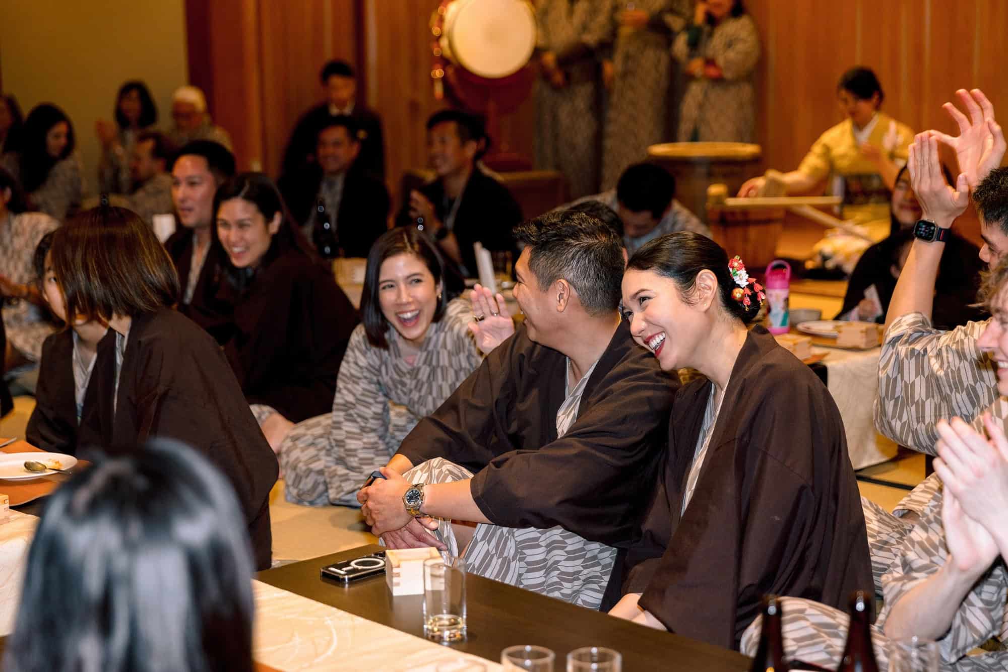 Guests in yukata laugh and clap at long tables in a wooden hall, wearing the ryokan's cotton robes to a wedding welcome dinner in Japan.