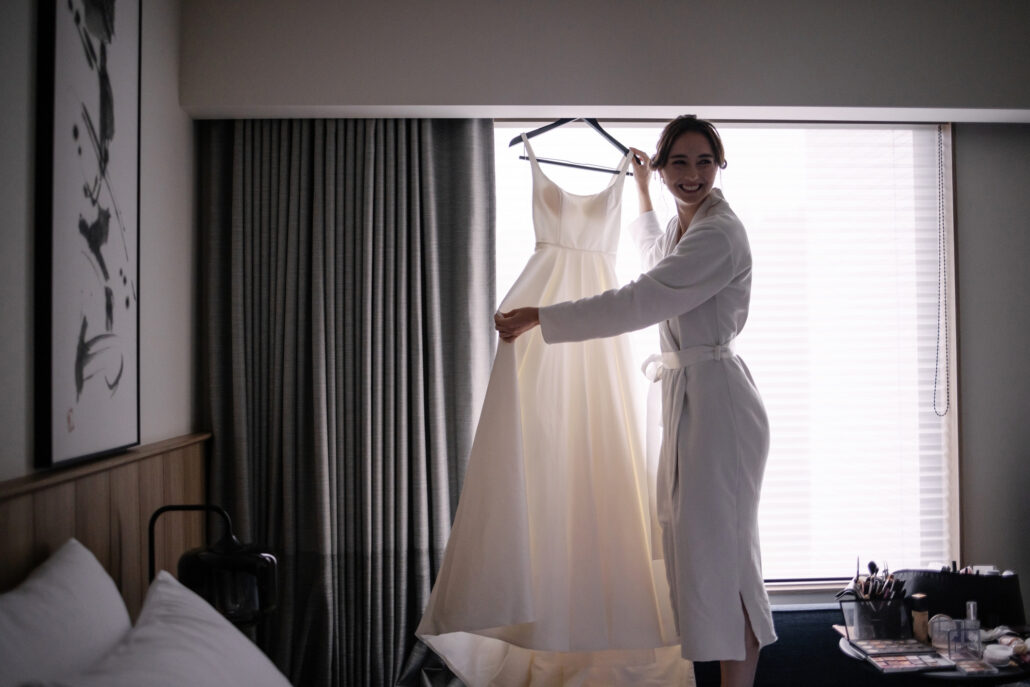 The bride in a robe lifts her wedding dress where it hangs at the window of her hotel room, a calligraphy scroll beside her.