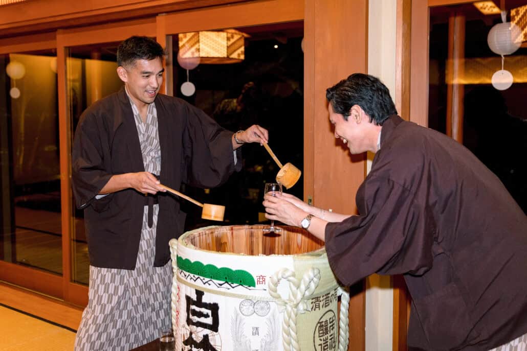 The groom ladles sake from the opened barrel into a guest's glass, both in yukata, at the welcome dinner the night before the wedding.
