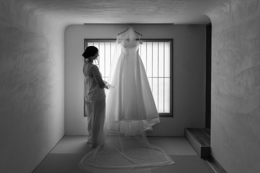In black and white, the bride stands at a barred window in a plaster room beside her wedding gown hanging from the frame.
