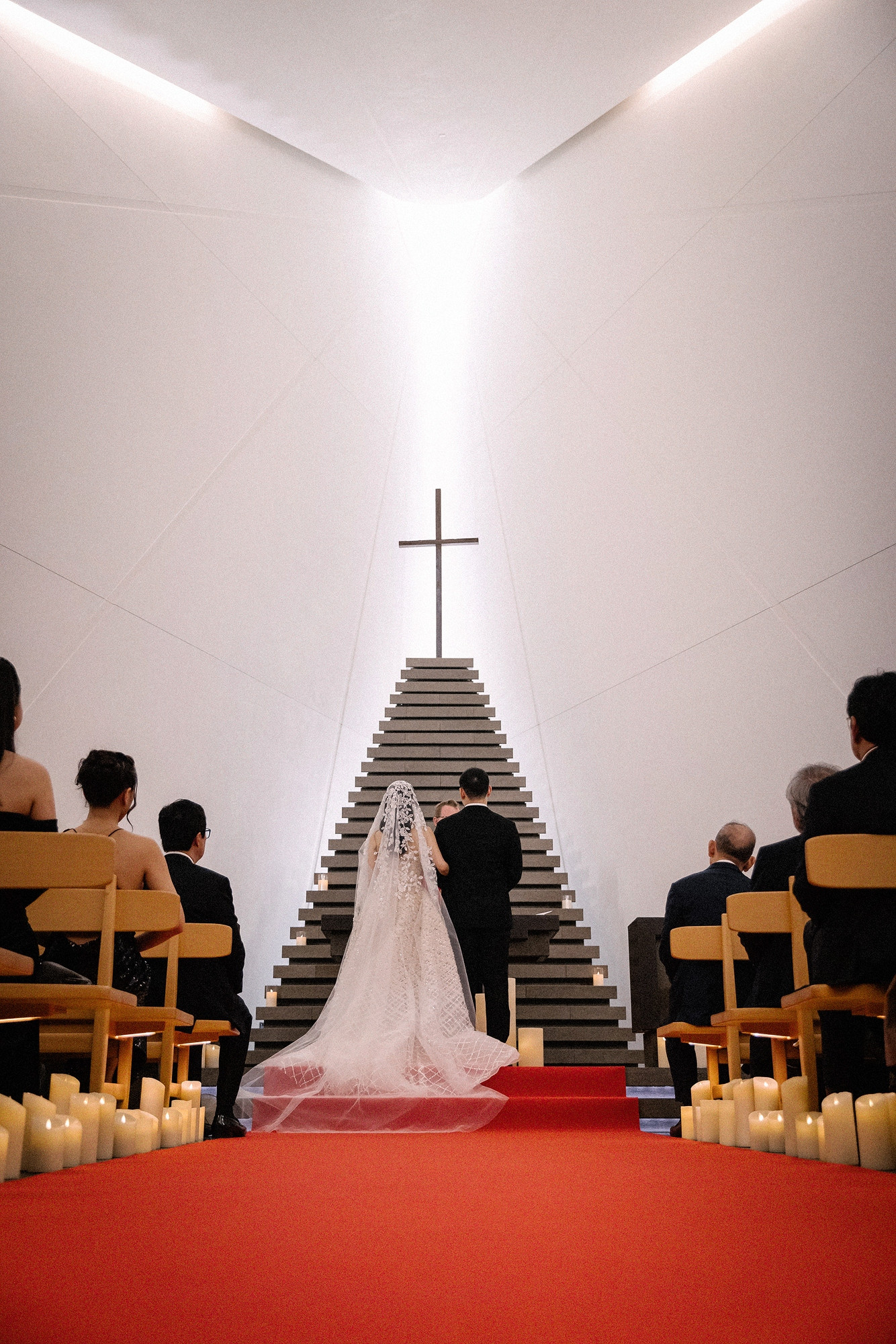 Light falls from above the cross onto a stepped stone form in the white Andaz Tokyo chapel as a couple marry, a hotel chapel wedding in Japan.