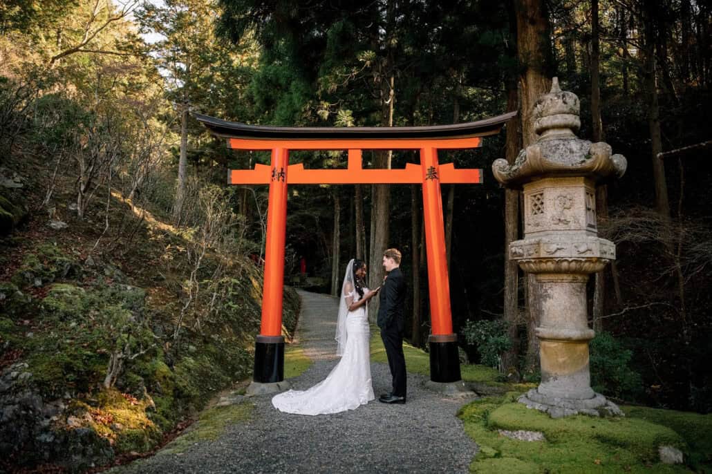 The bride and groom face each other beneath a vermilion torii beside a stone lantern and moss, a cedar forest behind their Kyoto elopement.
