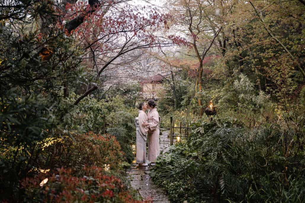 Two brides in pastel kimono kiss on a wet stone path lined with small lanterns in a lush Japanese garden with blossom beyond.