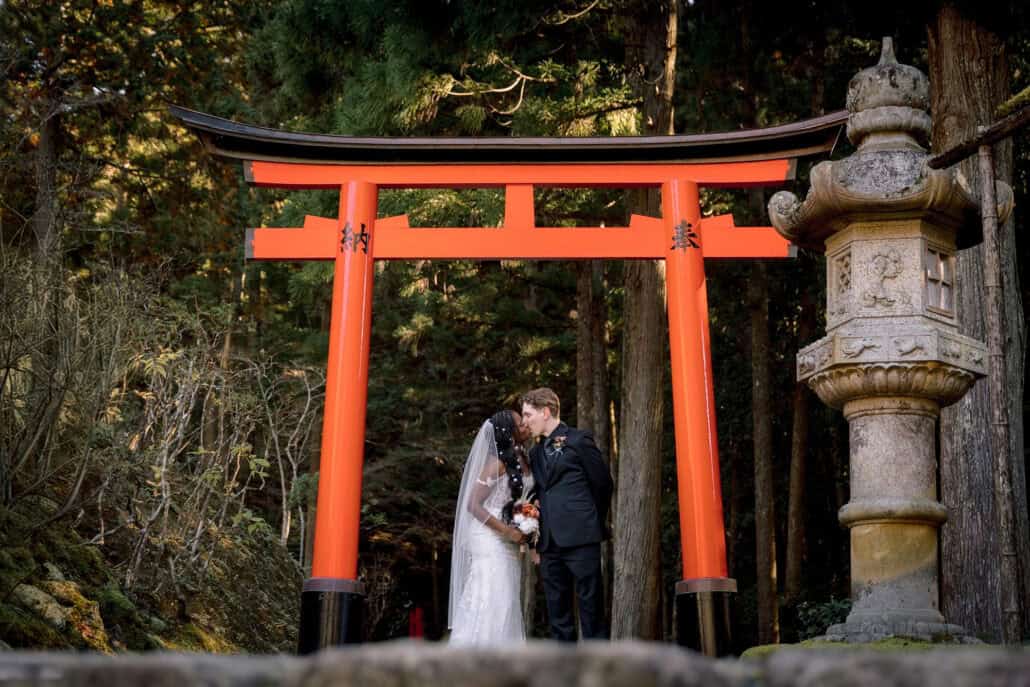 The bride and groom kiss beneath the vermilion torii, a stone lantern beside them and dark cedars behind, as the ceremony ends.