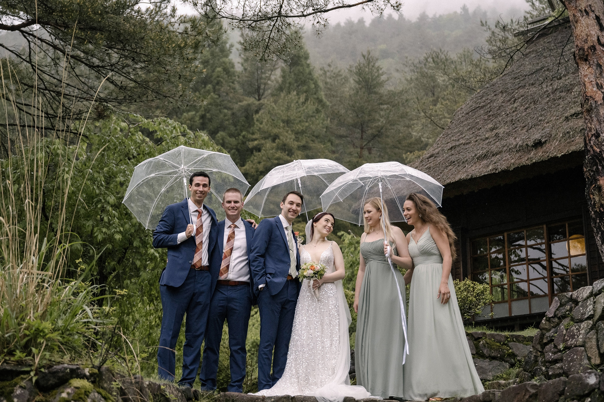 A couple and their four friends stand under clear umbrellas by a thatched house near Lake Saiko, where a rainy week moved a Mt Fuji wedding to a village built for wet weather.