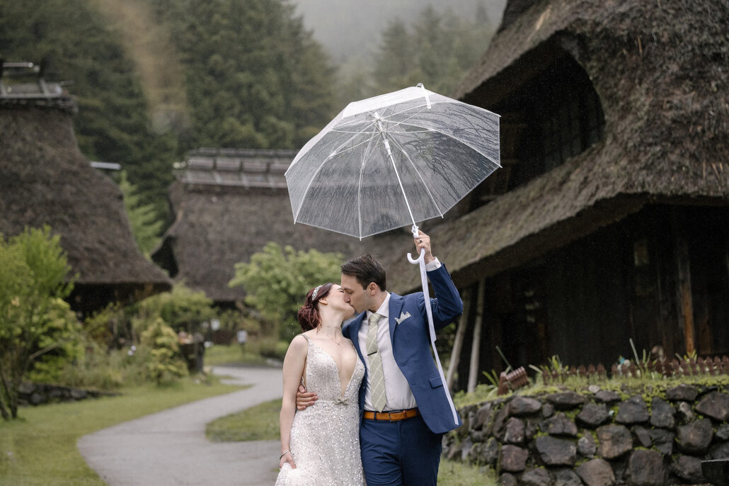 The couple kiss under a clear umbrella on a winding path through thatched houses at Saiko Iyashi no Sato Nenba in the rain.