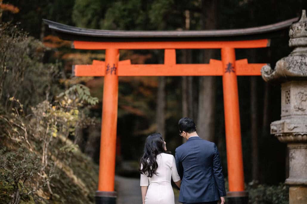 The couple walk hand in hand toward a vermilion torii and a tall stone lantern, cedars and moss all around, in Kyoto in autumn.