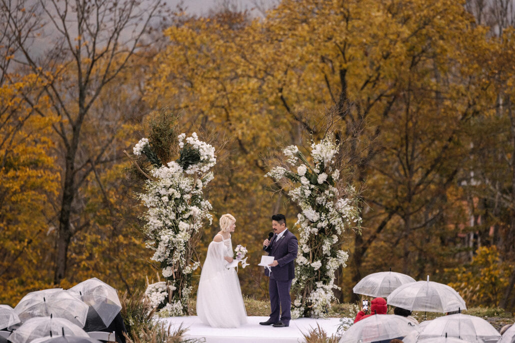 Guests under clear umbrellas watch the couple exchange vows between white flower columns at a wet, windy Niseko wedding.