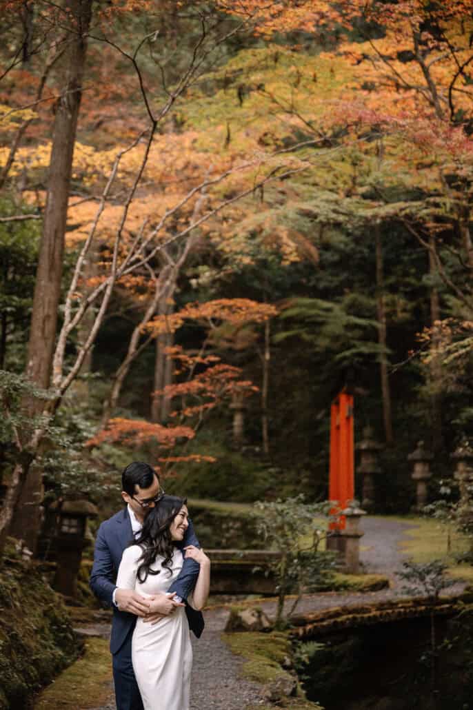 The groom holds the bride from behind on a mossy path in Kyoto, a vermilion torii and orange maples softly out of focus behind.