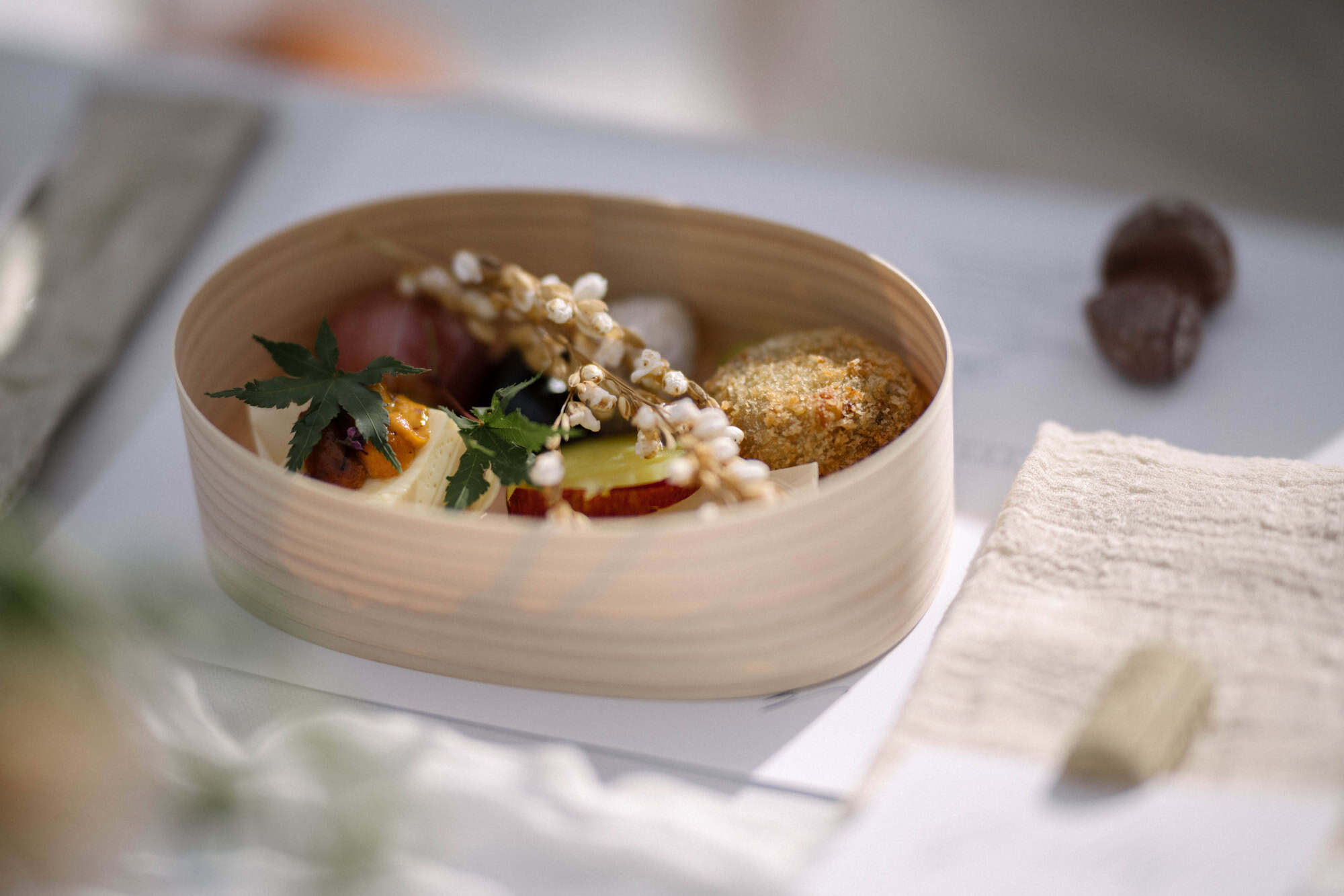 A bento lunch in a round bentwood box, with a croquette, vegetables and a maple leaf garnish, served at an autumn winery wedding in Japan.