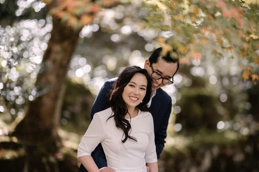 The groom holds the bride close in a sunlit Kyoto garden, the trunk of a great tree and autumn leaves glowing behind them.