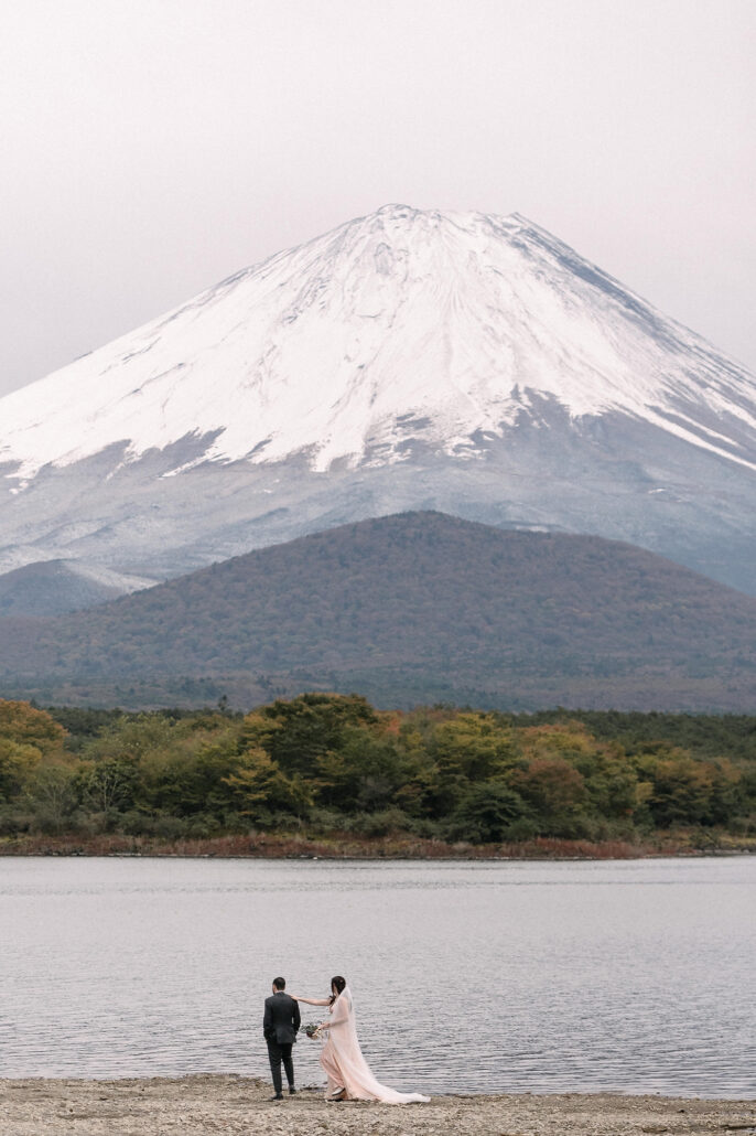 Snow-covered Mt Fuji fills the sky above Lake Shoji in late October, a couple small at the water's edge.