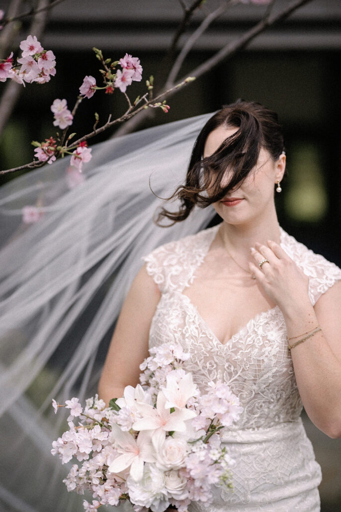 The wind blows the bride's hair across her face as she holds her bouquet beneath cherry blossom, her veil streaming beside her.
