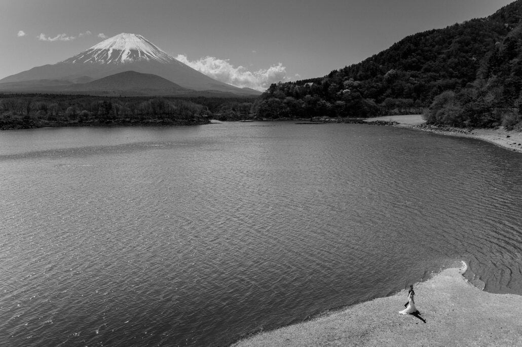 In black and white, Lake Shoji with Mt Fuji on the horizon and a bride small on a curve of the shore.