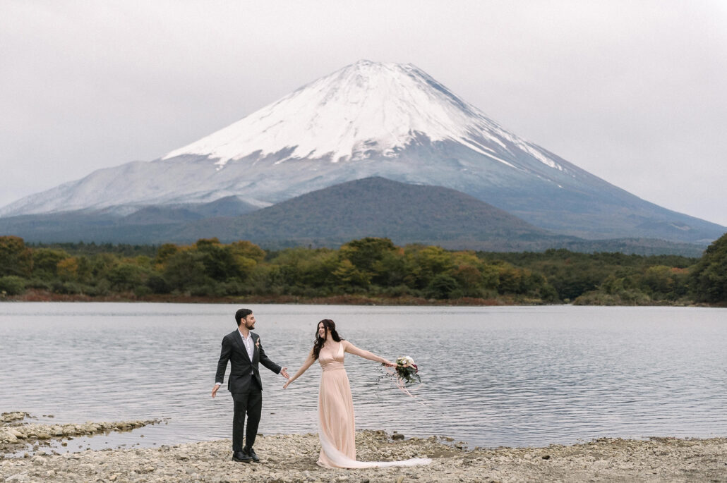 On the shore of Lake Shoji in late October, a couple walk hand in hand beneath Mt Fuji, white with autumn snow above the turning trees.