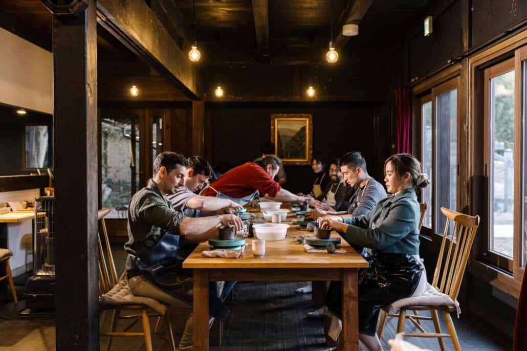 Friends bend over pottery wheels along one long table in an old house, making their own ceramics during a wedding in Japan.