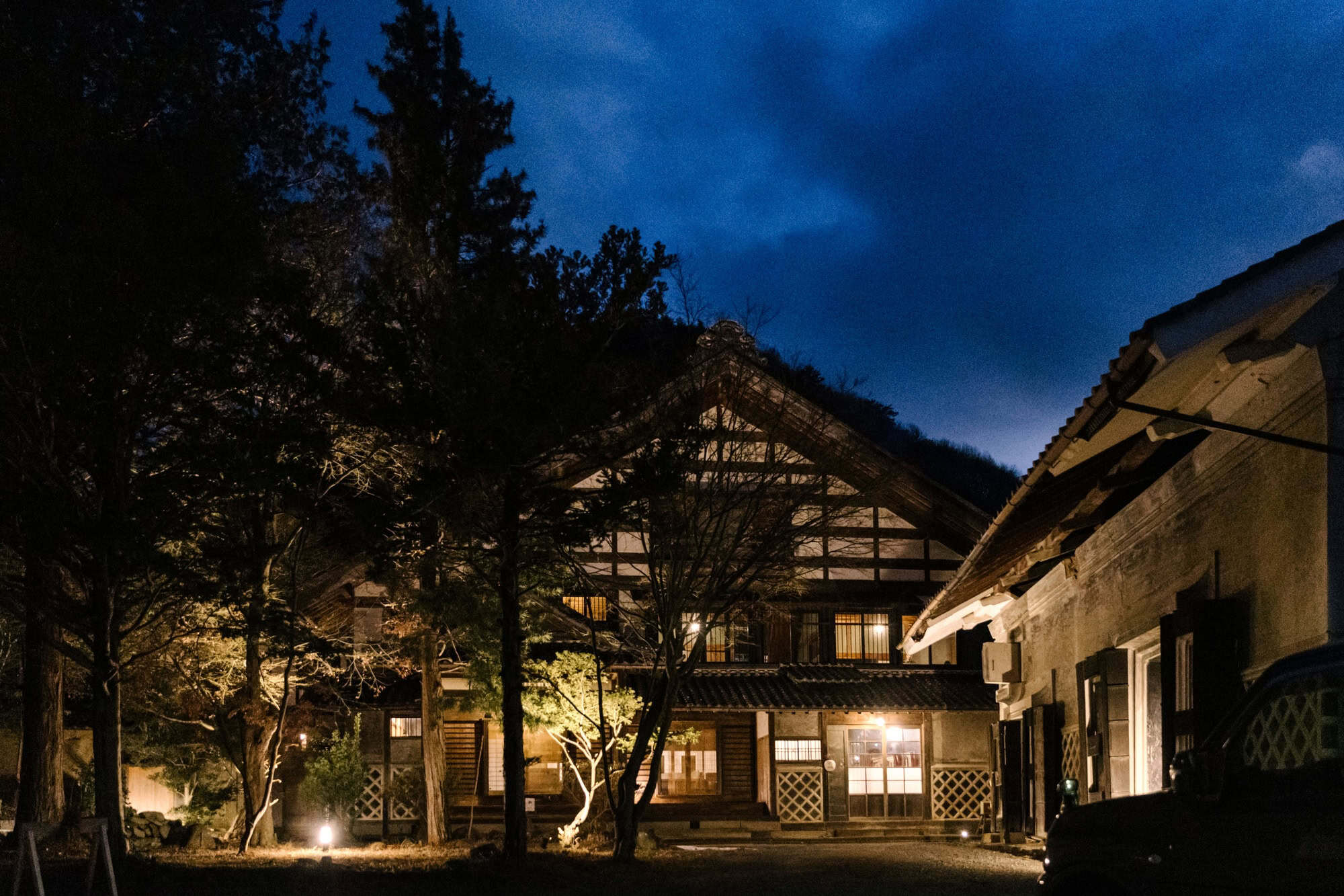 An old farmhouse glows from within at blue hour, its big gable and garden trees lit before the welcome dinner at a Nagano wedding venue.