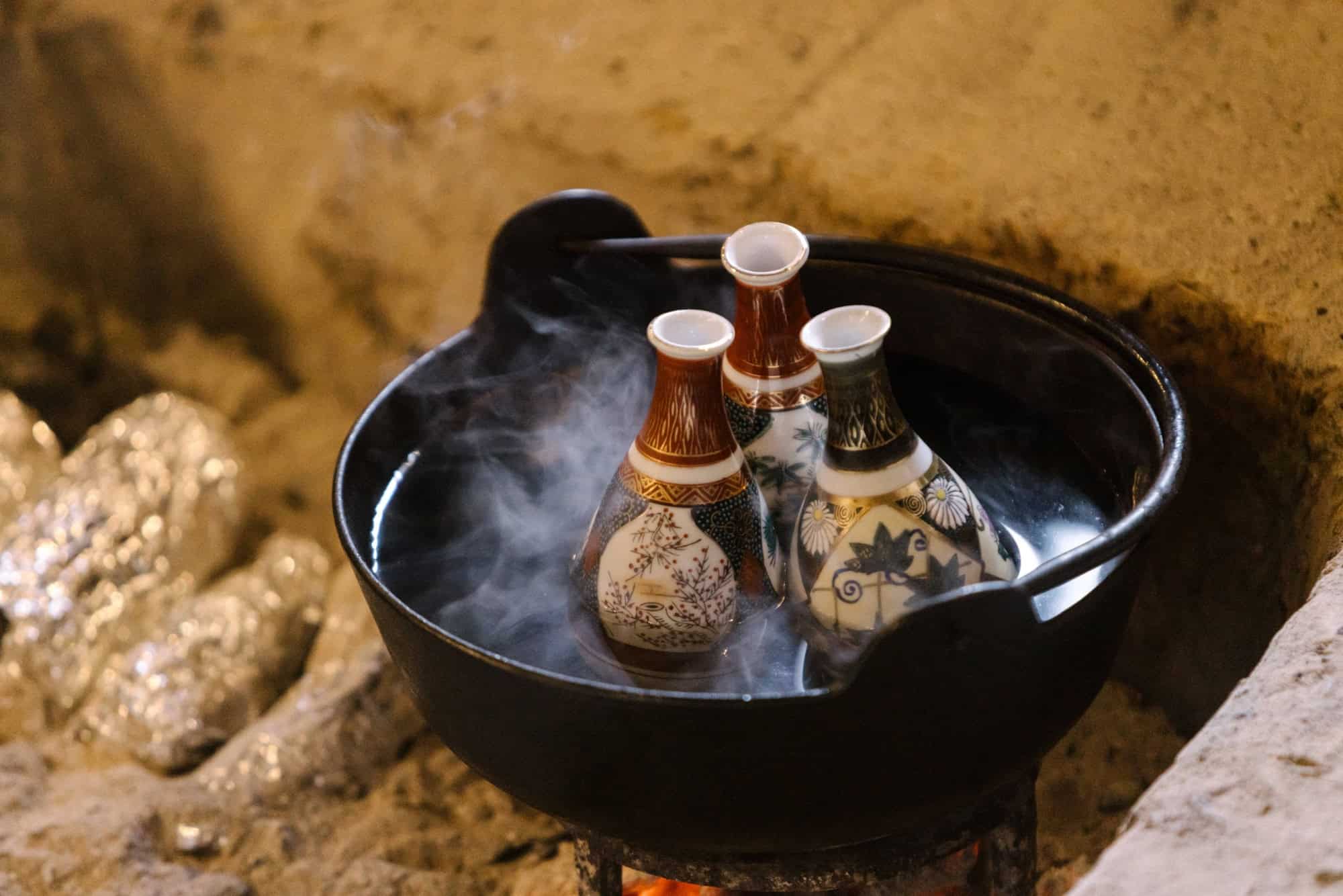 Sake flasks warm in an iron pot over the embers of an irori hearth, sweet potatoes in foil roasting in the ash, at a winter wedding in Nagano.