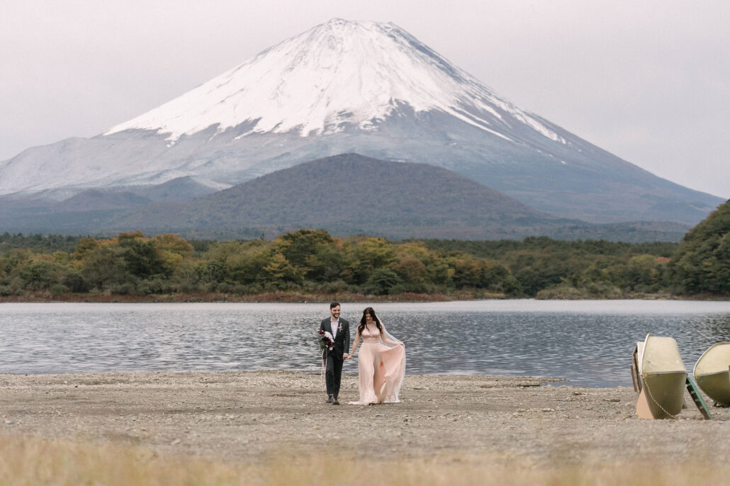 The couple walk across the stony shore of Lake Shoji beneath snow-capped Mt Fuji, with boats resting on the beach.