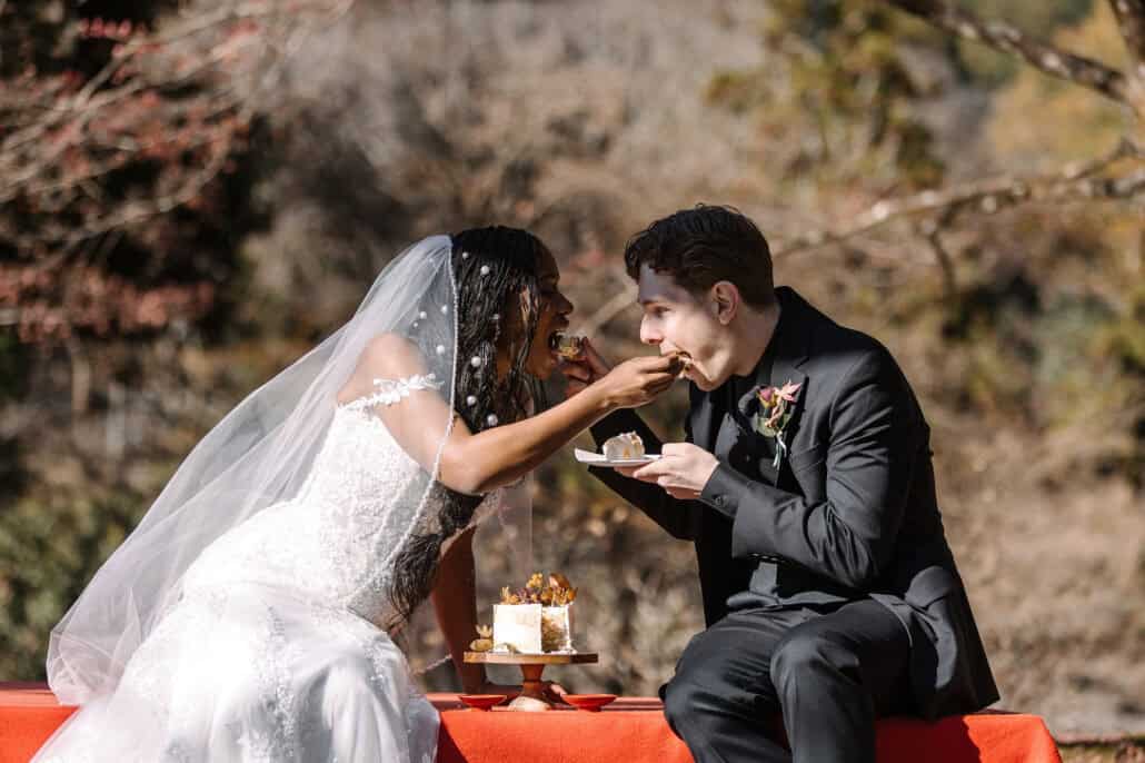 The bride feeds the groom a piece of wedding cake as they sit on a red felt bench in a Kyoto garden, autumn trees behind them.