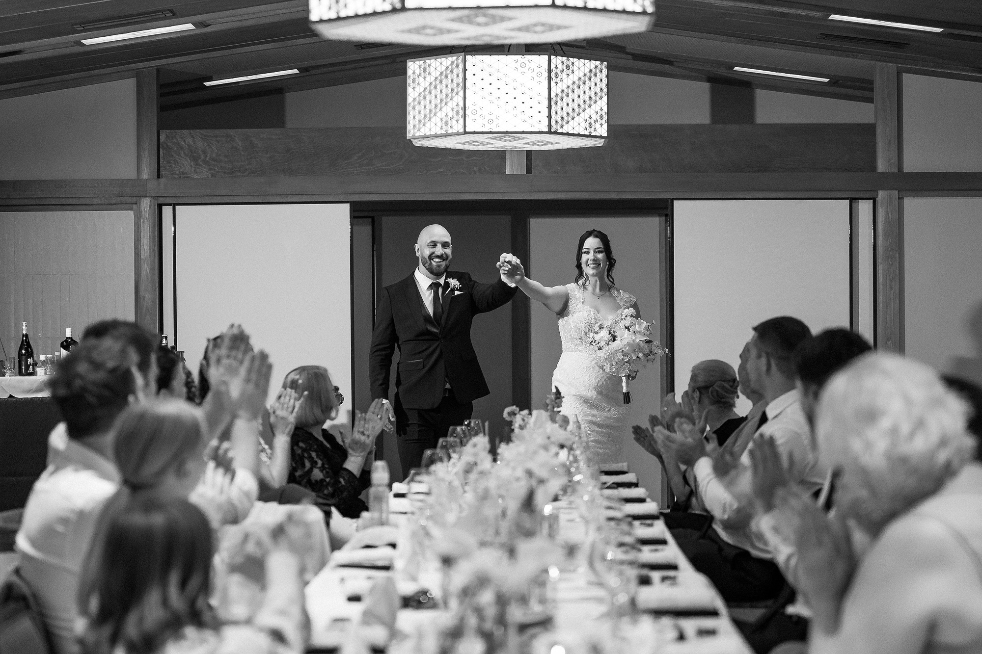 In black and white, the bride and groom raise their hands as they enter a ryokan dining room and guests applaud along one long dinner table.