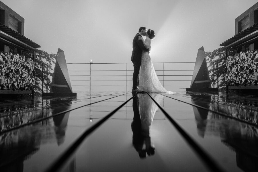 Black and white frame of the couple at the end of a wet timber deck in fog, their reflection running down the boards toward the camera.