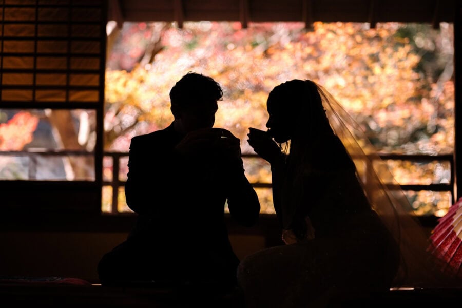The bride and groom drink matcha in silhouette in a tatami tea room, autumn maples glowing beyond the open window.