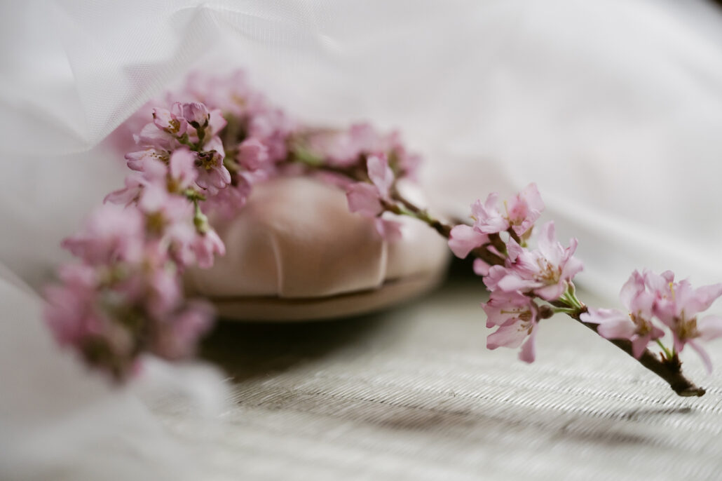 A sprig of pink cherry blossom rests across a soft pink bridal shoe on tatami, a detail from the wedding morning.