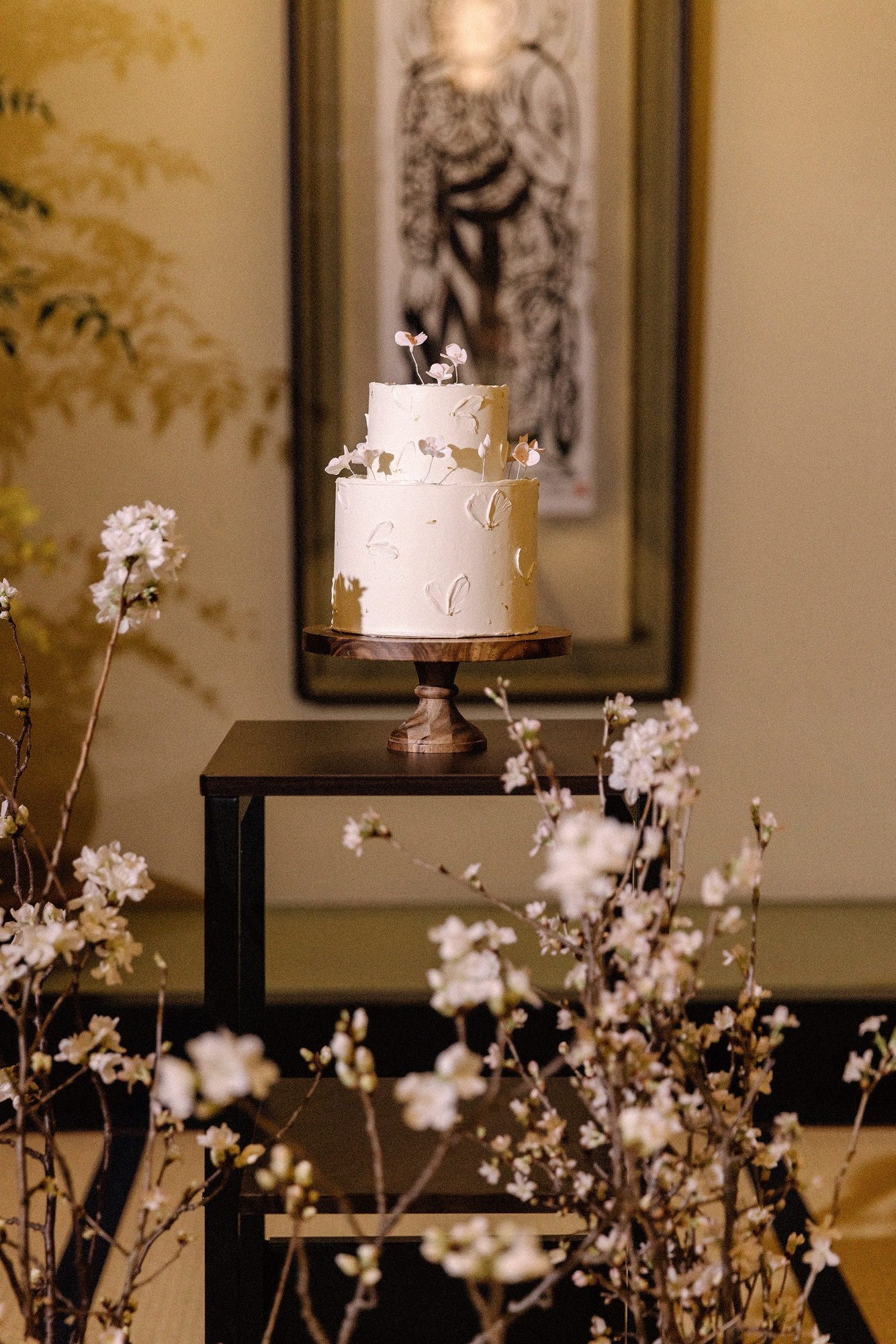 A white two-tier wedding cake on a wooden stand in a ryokan alcove, branches of white blossom in front, at a spring wedding in Japan.
