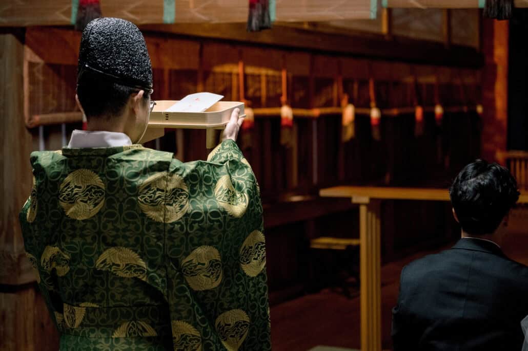 The priest in green robes raises a written prayer at the start of the wedding blessing as the groom sits in the hall.