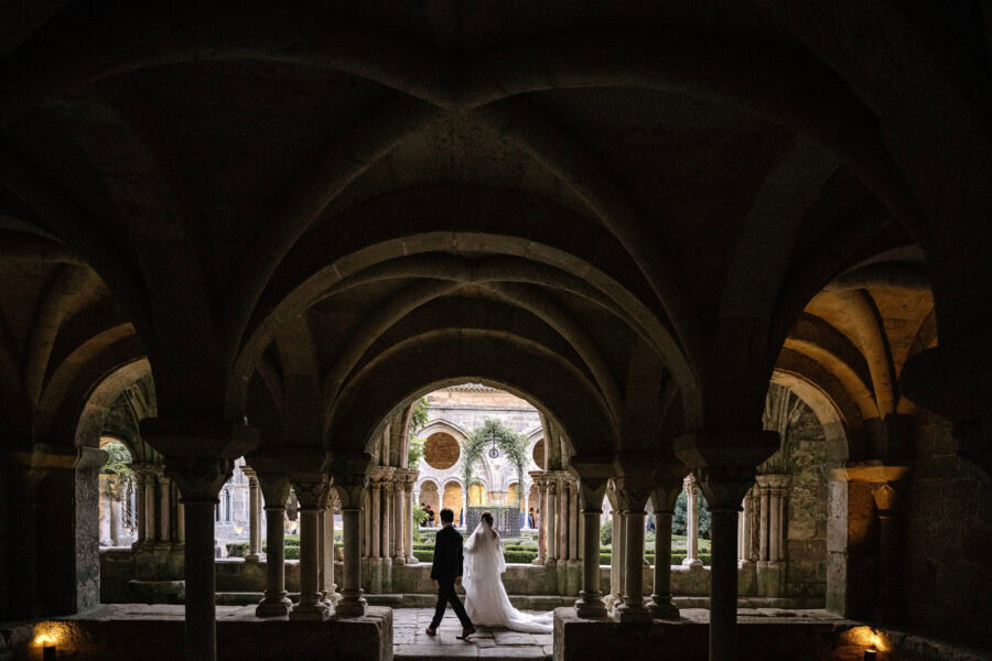 Under low ribbed vaults at the Abbaye de Fontfroide, the couple walks past columns into the light of the cloister garden beyond.