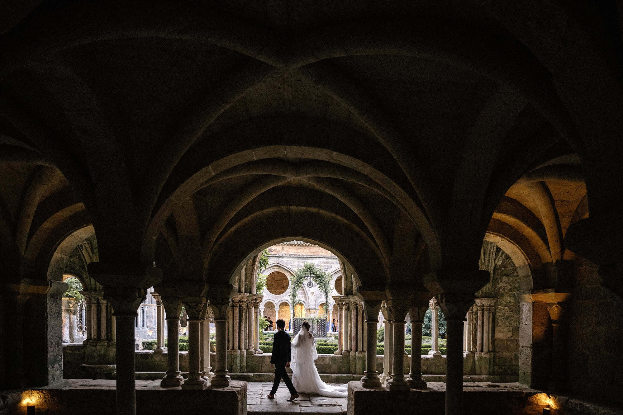 Under low ribbed vaults at the Abbaye de Fontfroide, the couple walks past columns into the light of the cloister garden beyond.