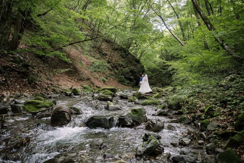Water runs over mossy rocks in a shaded forest gorge in Yamanashi, a bride a small white figure far up the river