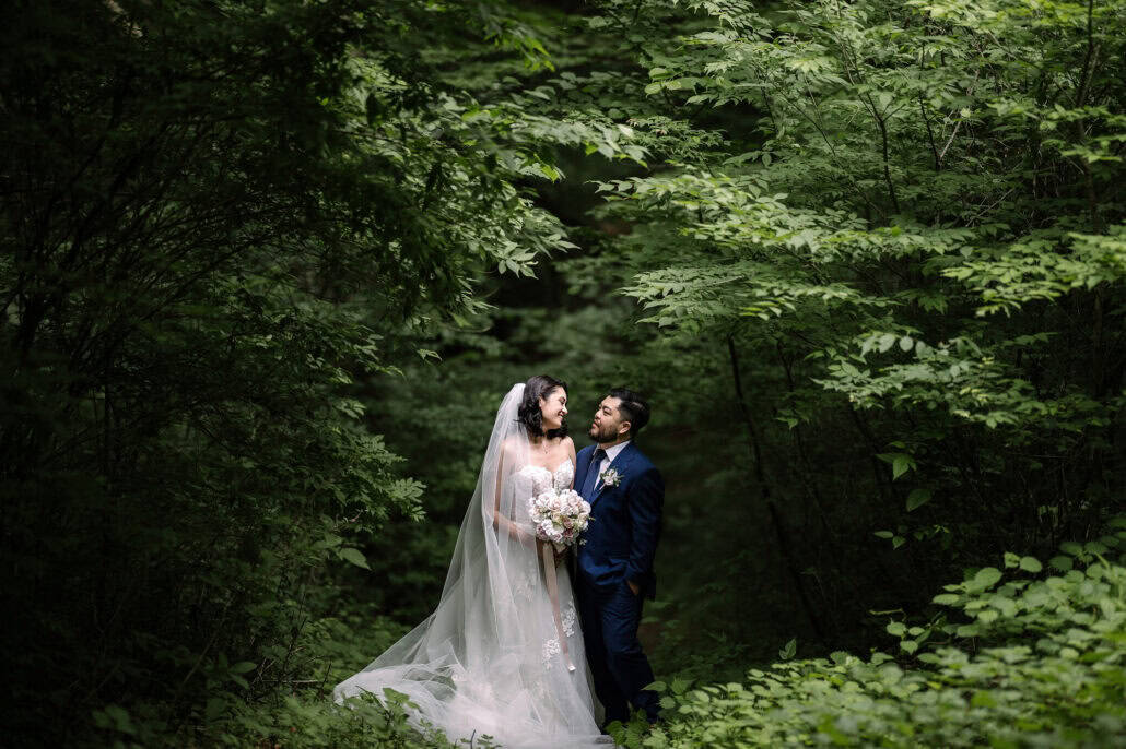 The bride and groom stand close in a forest of bright green leaves, her veil and bouquet pale against the dark trees.