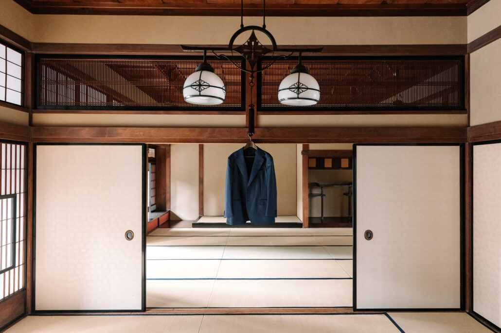 An empty tatami room in a traditional Japanese house, paper doors open and glass lamps hanging, a navy suit waiting on a hanger.