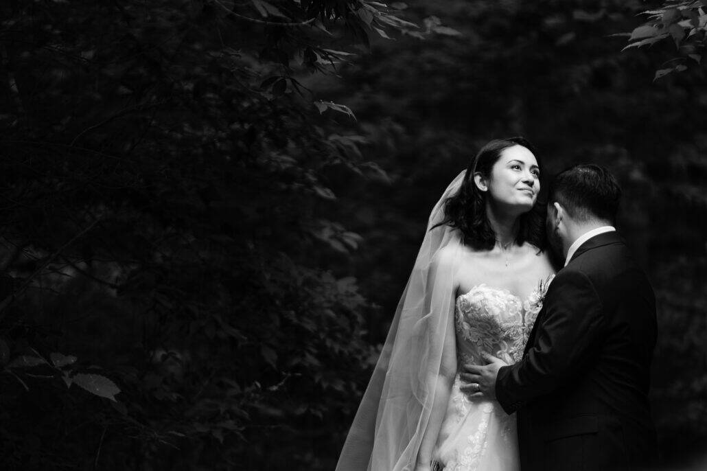 Black and white photograph of a bride looking up into the trees as the groom holds her close in a dark mountain forest in Japan