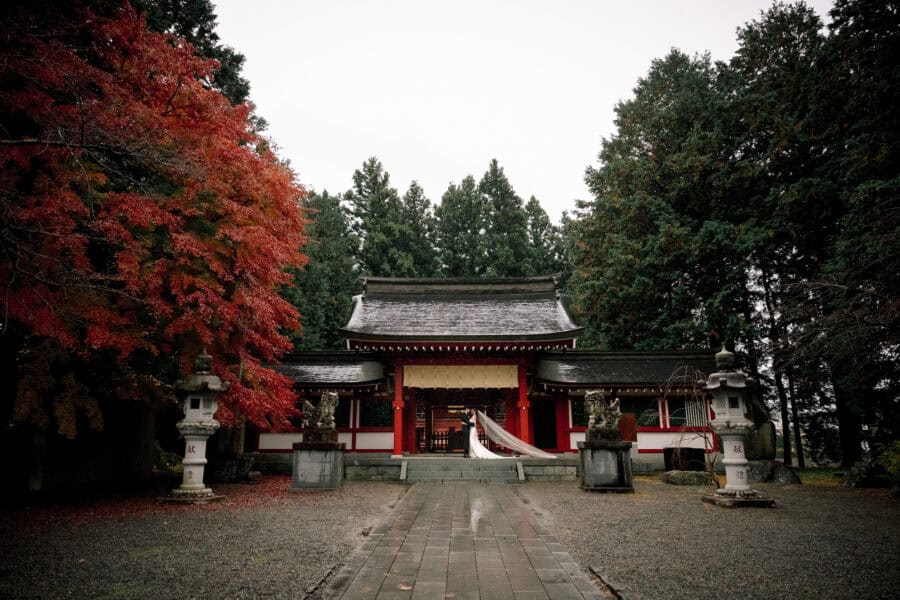 A wide view of the vermilion gate at the end of a wet gravel approach at Fuji Omuro Sengen Jinja, the bride's veil lifting in the wind.