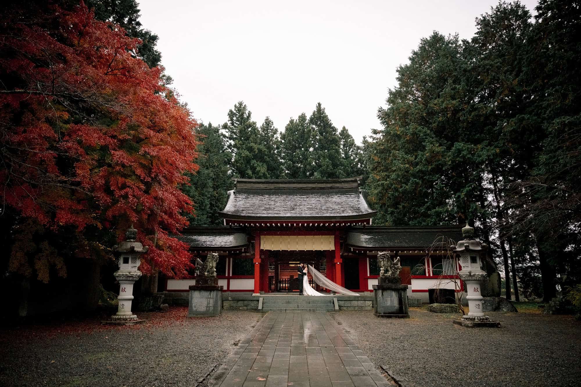 A wide view of the vermilion gate at the end of a wet gravel approach at Fuji Omuro Sengen Jinja, the bride's veil lifting in the wind.