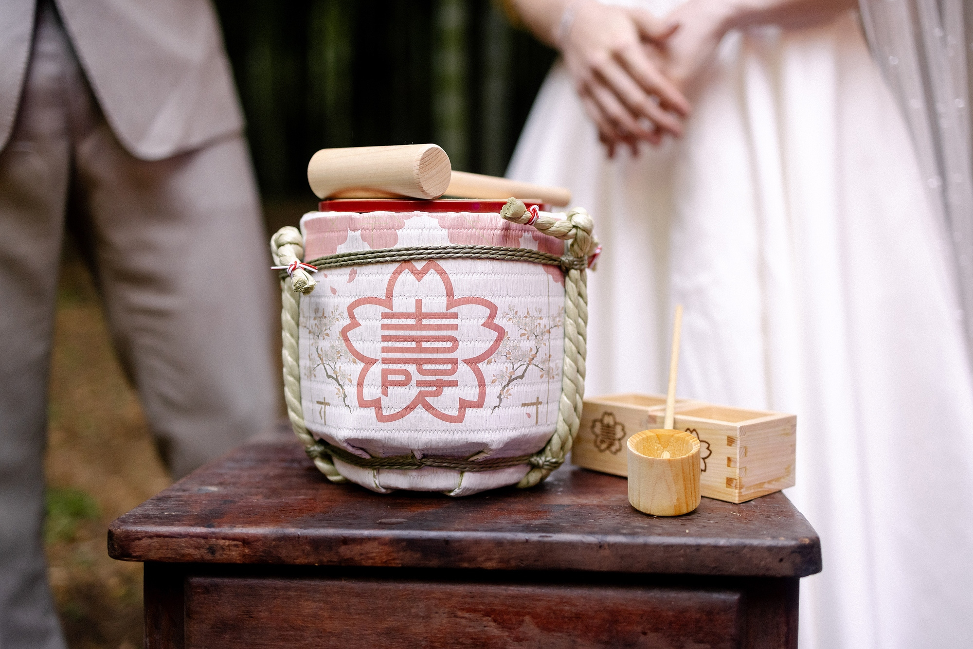 A small sake barrel wrapped in red and white sits on a wooden stand beside square wooden masu cups, ready for an elopement kagami biraki.