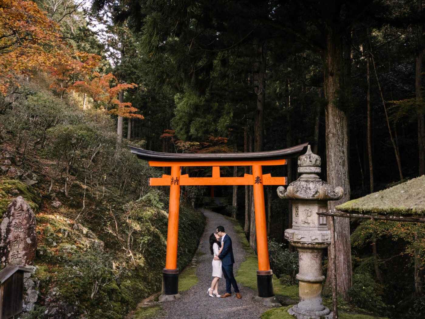 From above, the couple kiss beneath a red torii between a stone lantern and a moss-roofed pavilion at their private garden elopement in Kyoto.