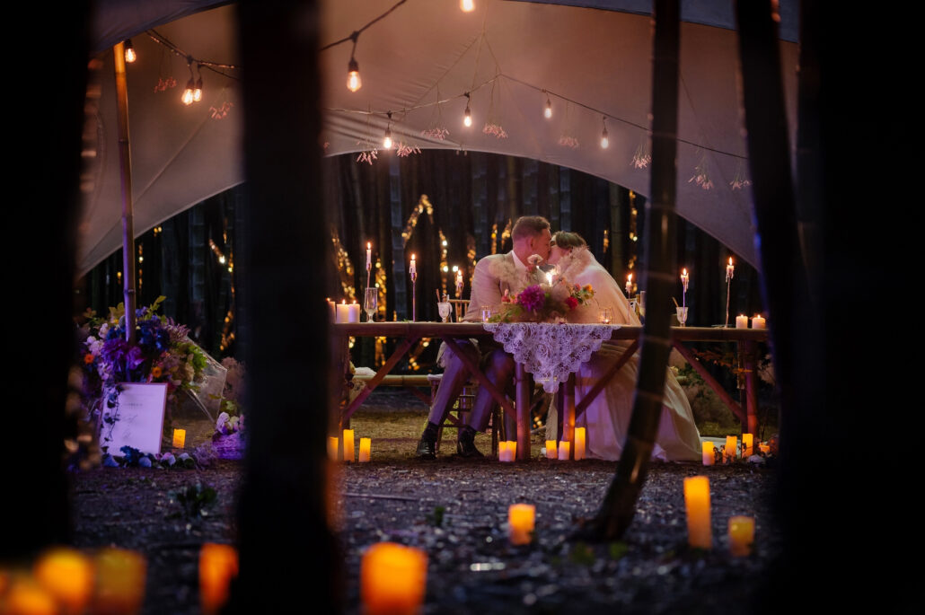 The white dinner canopy glows with candles and string lights through the dark bamboo grove at night.