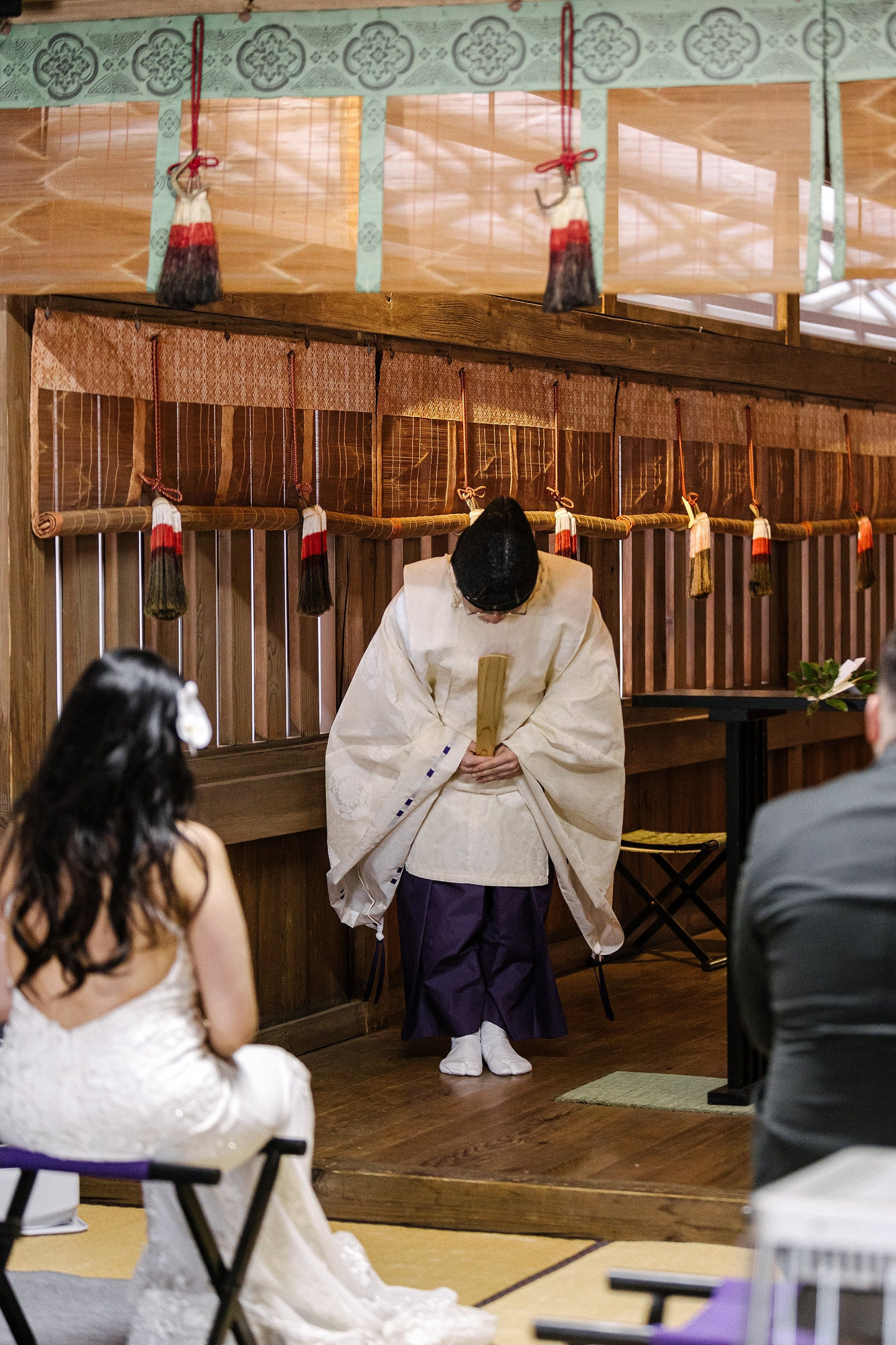 A priest in white and purple robes bows before the altar while a bride and groom sit in the hall, a Shinto shrine wedding blessing near Mt Fuji.