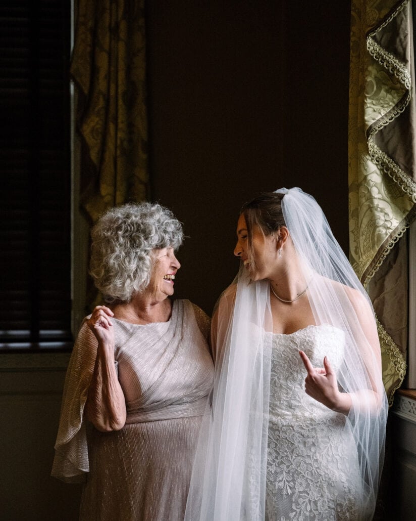 The bride laughs with her grandmother beside a tall window on her wedding day.