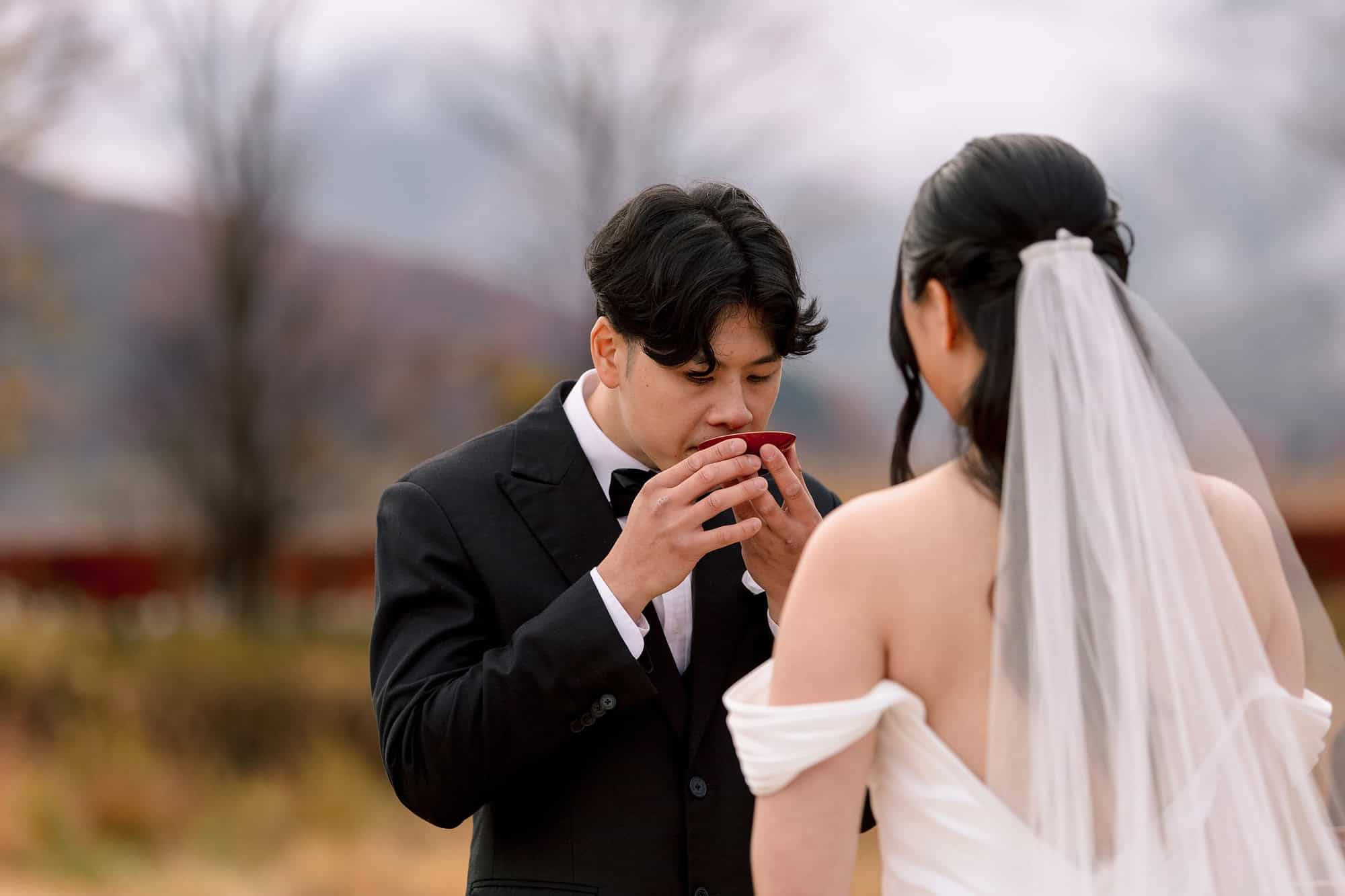 A groom sips sake from a red lacquer cup during san-san-kudo beside Lake Kawaguchiko as the bride watches, a lakeside ceremony near Mt Fuji.