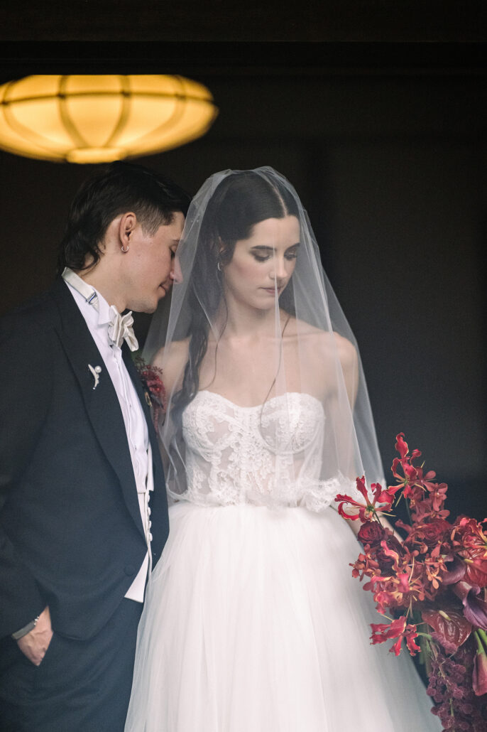 The groom leans in toward the bride, her red bouquet at her side, under a warm lamp.