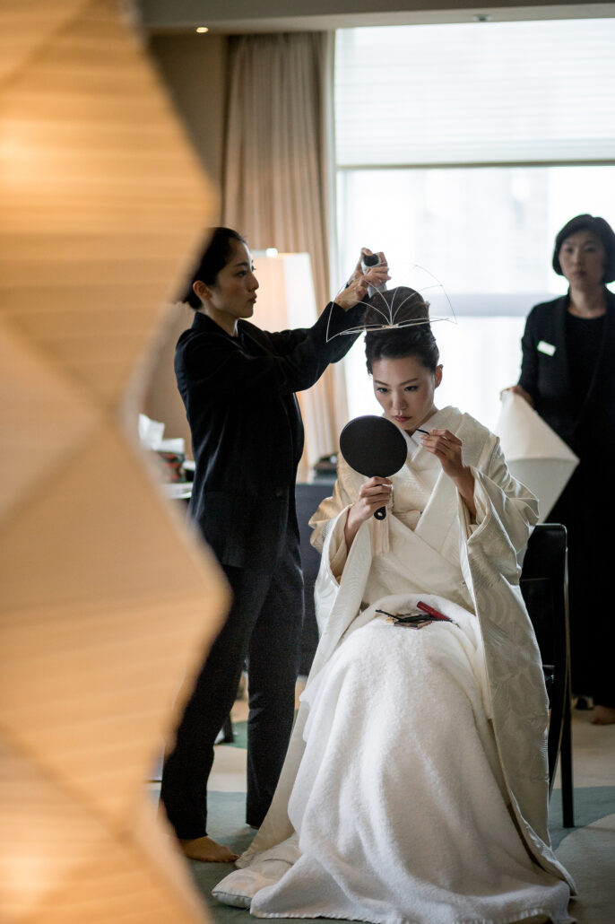 Two dressers fix a bride's hair as she sits in a white shiromuku kimono holding a hand mirror, on the morning of a Japanese wedding