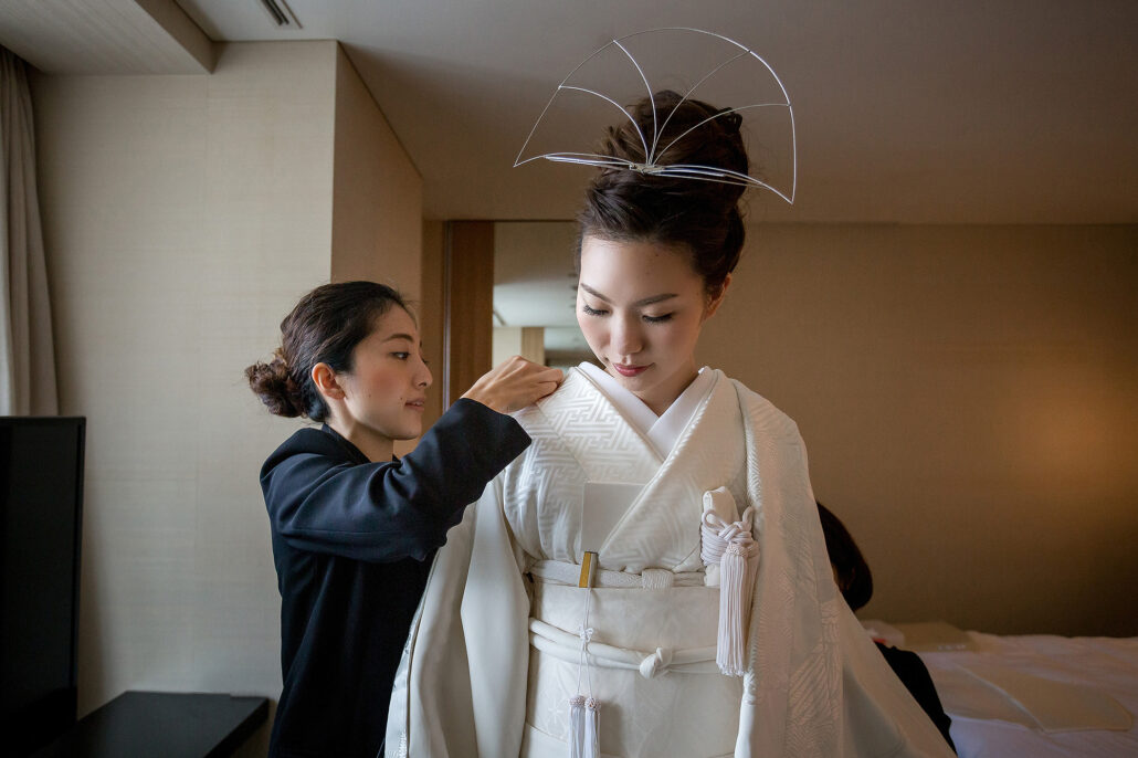 A kitsuke dresser smooths the collar of a bride's white shiromuku kimono, her hair set with a fine silver ornament before the hood goes on